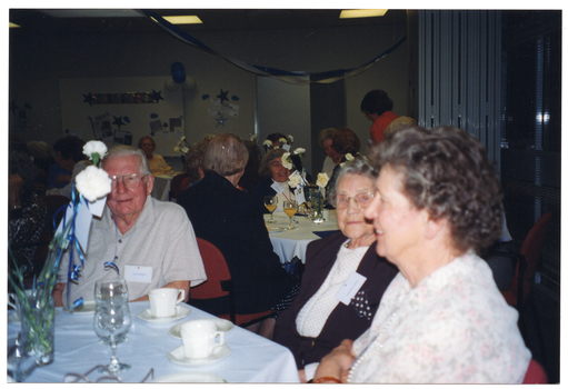 COL photo of several groups of people sitting around some tables. On the tables, there are vases of flowers, wine glasses, tea cups and saucers, and small plates. In the background, there are streamers and other decorations on the walls and hanging from the ceiling.