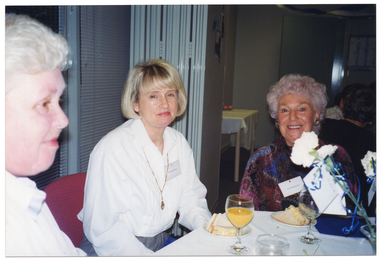 COL photo of three women sitting around a table. On the table, there are wine glasses, a vase of flowers, and small plates with sandwiches. In the background, there is another group of people (seated) and a small rectangular table (covered with a white tablecloth) against the wall, behind some open bi-fold doors.