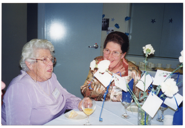 COL photo of two women sitting around a table. On the table, there are wine glasses, a vase of flowers, and small plates with sandwiches. To the left of the frame, the nose of a third person is only just evident. In the background, there are platters of sandwiches and cups and saucers on a table against the wall, which has decorations on it.