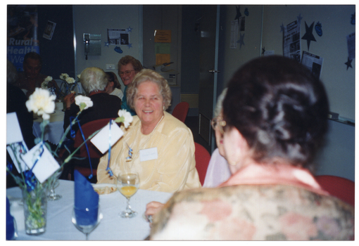 COL photo of three women (one partially obscured) sitting around a table. On the table, there are wine glasses, a vase of flowers, and small plates with sandwiches. In the background, other groups of people are also sitting at tables. There are posters and decorations on the walls.