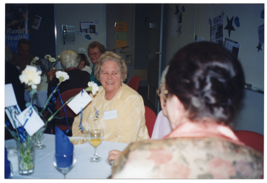 COL photo of three women (one partially obscured) sitting around a table. On the table, there are wine glasses, a vase of flowers, and small plates with sandwiches. In the background, other groups of people are also sitting at tables. There are posters and decorations on the walls.