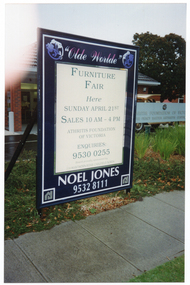 COL photo of a large advertising billboard affixed in the grassy verge in front of an office building. The text on the billboard is "Olde Worlde", "FURNITURE FAIR HERE SUNDAY APRIL 21ST SALES 10AM - 4PM ARTHRITIS FOUNDATION OF VICTORIA ENQUIRIES: 9530 0255", "Board proudly sponsored by 116 HAWTHORN ROAD CAULFIELD NORTH", "NOEL JONES 9232 8111". Beside and slightly behind the billboard, there is a long rectangular sign with the text, "ARTHRITIS FOUNDATION OF VICTORIA", followed by, "THE PERCY BAXTER ARTHRITIS CENTRE".
