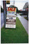 COL photo of a tall sign tied to an electricity pole on the nature strip. The sign consists of an A-frame sandwich board on the bottom, above to another sign above it. The text on the top sign is "SALE SUNDAY 21 April 10am - 4 pm". The text on the sandwich board is "CEDAR WORKS", [some text covered by an arrow drawn onto paper, taped on top), "ANTIQUE FURNITURE 22 PIN OAK CRESCENT PH. 376.3555 FACTORY PH.791.7044." There is also a bus stop sign affixed to the power pole.