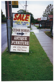 COL photo of a tall sign tied to an electricity pole on the nature strip. The sign consists of an A-frame sandwich board on the bottom, above to another sign above it. The text on the top sign is "SALE SUNDAY 21 April 10am - 4 pm". The text on the sandwich board is "CEDAR WORKS", [some text covered by an arrow drawn onto paper, taped on top), "ANTIQUE FURNITURE 22 PIN OAK CRESCENT PH. 376.3555 FACTORY PH.791.7044." There is also a bus stop sign affixed to the power pole.