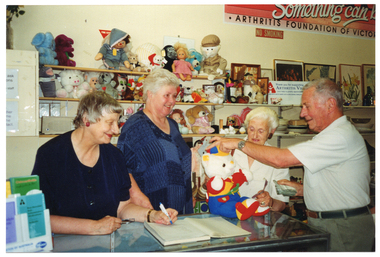 COL photo of four people inside a shop. Three women are standing behind the shop counter, while a man stands on the other side. One woman (to the left of the frame) is holding a pen, poised as if about to write in a ledger book. In her other hand, she is holding a pair of spectacles. Another woman (to the right of the frame) is holding the back of a teddy bear, which is sitting upright on the countertop. The man has one hand placed on top of the teddy bear's head. He is holding a handkerchief with his other hand. The woman in the centre is looking at the man. On the left of the counter, there are various Arthritis Victoria brochures on a small display stand. On the right, there is a plastic ruler across the corner of the counter. Directly behind the women are some shelves with soft toys and dolls. Beside that are more shelves containing crockery, bric-a-brac, paintings and prints. On the top shelf, there is a white rectangular sign that says, "Thank you for supporting ARTHRITIS VICTORIA your help is greatly appreciated". There is another line of text below, which is illegible. Beside the text, 'Arthritis Victoria' is the organisation's logo. On the wall above the shelves is a large rectangular sign (partially obscured), which says "Something can b... ARTHRITIS FOUNDATION OF VICTO...". Below that is a NO SMOKING sign.