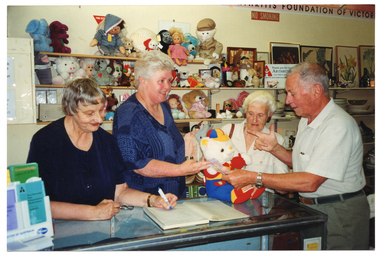 COL photo of four people inside a shop. Three women are standing behind the shop counter, while a man stands on the other side. One woman (to the left of the frame) is holding a pen, poised as if about to write in a ledger book. In her other hand, she is holding a pair of spectacles. Another woman (to the right of the frame) is holding the back of a teddy bear, which is sitting upright on the countertop. The man hands over a $5 note to the woman standing in the centre. On the left of the counter, there are various brochures on a small display stand. On the right, there is a plastic ruler along the front edge of the counter. Directly behind the women are some shelves with soft toys and dolls. Beside that are more shelves, with crockery, bric-a-brac, paintings and prints. On the top shelf, there is a white rectangular sign (partially obscured) that says, "Thank you for supporting ARTHRITIS VICTORIA your help is greatly appreciated". There is another line of text below, which is illegible. Beside the text 'Arthritis Victoria' is the organisation's logo. On the wall above the shelves is a large sign (mostly obscured). Below that is a NO SMOKING sign.