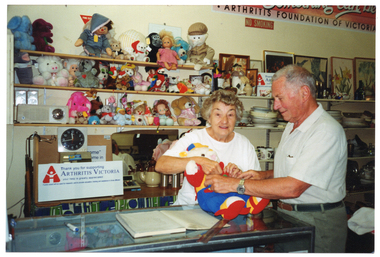 COL photo of two people inside a shop. One woman is standing behind the shop counter, while a man stands on the other side. They are both adjusting clothing on a teddy bear, which is sitting on the front counter. On the counter, there is a pen and large ledger book with a plastic ruler across the top corner. On the wall behind them are some shelves with soft toys and dolls, a radio and a clock. Beside that are more shelves, with crockery, bric-a-brac, paintings and prints. On the top shelf, there is a white rectangular sign Arthritis Victoria sign, (partially obscured). On benchtop behind them, there is another sign, which is exactly the same. It says, "Thank you for supporting ARTHRITIS VICTORIA your help is greatly appreciated", and "Funds raided will be used for research, and to provide education, training, and assistance to those affected". Beside the text 'Arthritis Victoria' is the organisation's logo. On the wall above the shelves is a large rectangular sign (partially obscured), which says "ARTHRITIS FOUNDATION OF VICTORIA" at the bottom. Below that is a NO SMOKING sign.