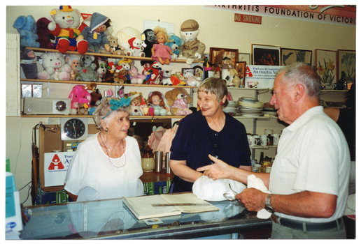 COL photo of three people inside a shop. Two women are standing behind the shop counter, while a man stands on the other side. They are all chatting to one another and smiling. The man is holding a pair of spectacles and something white (indiscernable) in one hand. On top of the counter, there is a ledger book with a plastic ruler across the top, a plastic bag with something bulky inside, and some brochures on a small display stand (partially obscured). On the wall behind them, there are some shelves with soft toys and dolls, a radio and a clock. Beside that are more shelves, with crockery, bric-a-brac, paintings and prints. On the top shelf, there is a white rectangular sign (partially obscured) that says, "Thank you for supporting ARTHRITIS VICTORIA your help is greatly appreciated". There is another line of text below, which is illegible. Beside the text, 'Arthritis Victoria' is the organisation's logo. On the wall above the shelves is a large rectangular sign (partially obscured), which says "ARTHRITIS FOUNDATION". Below that is a NO SMOKING sign. There is a similar sign on the benchtop or shelf behind the women.