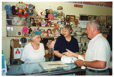 COL photo of three people inside a shop. Two women are standing behind the shop counter, while a man stands on the other side. They are all chatting to one another and smiling. The man is holding a pair of spectacles and something white (indiscernable) in one hand. On top of the counter, there is a ledger book with a plastic ruler across the top, a plastic bag with something bulky inside, and some brochures on a small display stand (partially obscured). On the wall behind them, there are some shelves with soft toys and dolls, a radio and a clock. Beside that are more shelves, with crockery, bric-a-brac, paintings and prints. On the top shelf, there is a white rectangular sign (partially obscured) that says, "Thank you for supporting ARTHRITIS VICTORIA your help is greatly appreciated". There is another line of text below, which is illegible. Beside the text, 'Arthritis Victoria' is the organisation's logo. On the wall above the shelves is a large rectangular sign (partially obscured), which says "ARTHRITIS FOUNDATION". Below that is a NO SMOKING sign. There is a similar sign on the benchtop or shelf behind the women.