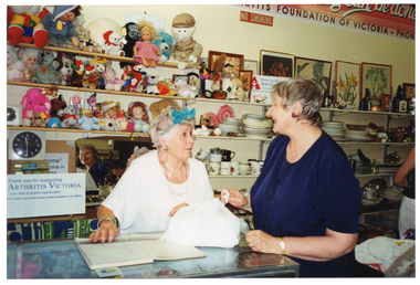 COL photo of two women inside a shop. They are standing behind and around the shop counter, chatting. On top of the counter, there is a ledger book with a plastic ruler across the top, a plastic bag with something bulky inside, and some brochures on a small display stand (partially obscured). On the wall behind them, there are some shelves with soft toys and dolls, a radio, a clock, and an Arthritis Victoria sign. The sign says, "Thank you for supporting ARTHRITIS VICTORIA your help is greatly appreciated". There is another line of text below, which is illegible. Beside the text 'Arthritis Victoria' is the organisation's logo. Beside that shelf are more shelves, with crockery, bric-a-brac, paintings and prints. On the top shelf, there is the same Arthritis Victoria sign with blue text and a red a logo. On the wall above the shelves is a large rectangular sign (partially obscured), which says "ARTHRITIS FOUNDATION OF VICTORIA. PHONE...". Below that is a NO SMOKING sign.