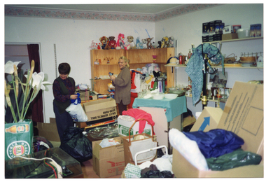 COL photo of two women inside a shop. They are sorting through bags and boxes of donations in a crowded room. Behind them is a bookcase with soft toys across the top, and other shelves with various items, including some boxed items. In front of the shelves is a hat stand, with a fabric cloth across the top. In the foreground, there is a shopping trolley, some artificial flowers in a box, a record player/turntable, and more boxes.