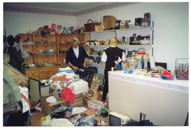 COL photo of two women inside a shop. They are sorting through bags and boxes of donations in a crowded room. Behind them is a large dresser, with drawers at the bottom and a hutch with shelves at the top. There are soft toys across the top shelf, and various other items on the shelves below. On the perpendicular wall, there are more shelves with various items, including cane baskets, a large straw hat, bric-a-brac, crockery, glassware, plastic kitchen storage containers, and some boxed items on the top shelf. The floor is crowded with boxes, baskets, and other miscellaneous items - including a portalbe television set. In the foreground, there are piles of clothing or fabric.