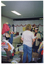 COL photo of two women inside a shop. They are sorting through bags and boxes of donations in a crowded room. One of them holds some pieces of black netting and white fabric on her head. Behind them is a counter with various items on top. The floor is crowded with boxes and trolleys full of clothing and other miscellaneous items.