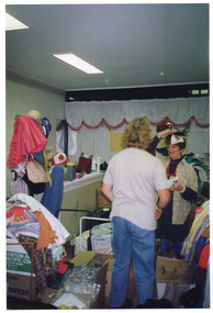 COL photo of two women inside a shop. They are sorting through bags and boxes of donations in a crowded room. One of them holds some pieces of black netting and white fabric on her head. Behind them is a counter with various items on top. The floor is crowded with boxes and trolleys full of clothing and other miscellaneous items.
