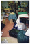 COL photo of a woman inside a crowded shop. She is sorting through bags and boxes of donations. She is holding up a gold decoration. On one side of the room, there is a wooden table. On top of the table are some artificial flowers in a box, some mugs, and some clothes. In the centre of the room, there is a small low table covered with a blue tablecloth. On top of the table is a plastic container of cut sandwiches, two glasses of milk, and two bananas.