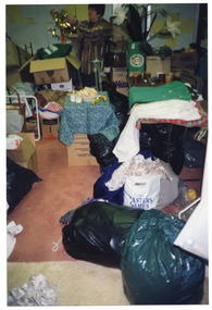 COL photo of a woman inside a crowded shop. She is sorting through bags and boxes of donations. She is holding up a gold decoration. On one side of the room, there is a wooden table. On top of the table are some artificial flowers in a box, some mugs, and some clothes. In the centre of the room, there is a small low table covered with a blue tablecloth. On top of the table is a plastic container of cut sandwiches, two glasses of milk, and two bananas.