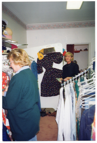 COL photo of two women inside a shop. One woman is tidying up some clothes which are hanging from racks, while another woman looks on from near a doorway. There is a dress, scarf, and handbag affixed to a partition, on display. In front of the partition are several pairs of shoes, lined up on the floor.