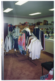 COL photo of the interior of a shop. There are several clothes racks in the centre of the room, with clothes hanging neatly from them. In the background, there is a counter, bookshelves, and some wall-mounted shelves. Various donated items have been arranged neatly on the shelving, including soft toys, baskets, some boxed items, kitchenware, and bric-a-brac.