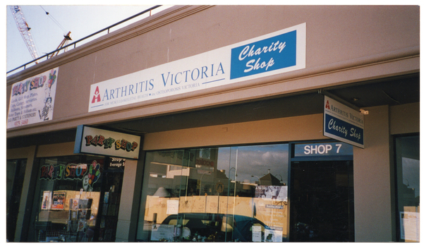 COL medium close up photo of two shops. Above the shop front is a sign on the veranda roof which displays the Arthritis Victoria logo and the text, "ARTHRITIS VICTORIA Charity Shop FOR MUSCULO-SKELETAL HEALTH . inc OSTEOPOROSIS VICTORIA". A similar sign hangs near the shop door from beneath the veranda ceiling. Above the door is a sign with the text, "SHOP 7". In the front window, there are posters and information sheets, including a Tai Chi poster, as well as a dinner plate and some bric-a-brac on display. The adjoining shop has three signs with the text heading, "Party Shop". One sign hangs from the veranda ceiling; the second is painted on the shop's front window with the addition of a cartoon drawing of a clown holding some balloons; and a third larger sign with additional information and the same clown cartoon is affixed to the top of the building above the shop. On the roof of the building, part of a crane is evident.