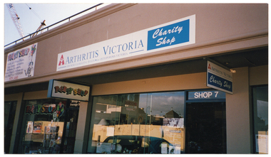 COL medium close up photo of two shops. Above the shop front is a sign on the veranda roof which displays the Arthritis Victoria logo and the text, "ARTHRITIS VICTORIA Charity Shop FOR MUSCULO-SKELETAL HEALTH . inc OSTEOPOROSIS VICTORIA". A similar sign hangs near the shop door from beneath the veranda ceiling. Above the door is a sign with the text, "SHOP 7". In the front window, there are posters and information sheets, including a Tai Chi poster, as well as a dinner plate and some bric-a-brac on display. The adjoining shop has three signs with the text heading, "Party Shop". One sign hangs from the veranda ceiling; the second is painted on the shop's front window with the addition of a cartoon drawing of a clown holding some balloons; and a third larger sign with additional information and the same clown cartoon is affixed to the top of the building above the shop. On the roof of the building, part of a crane is evident.