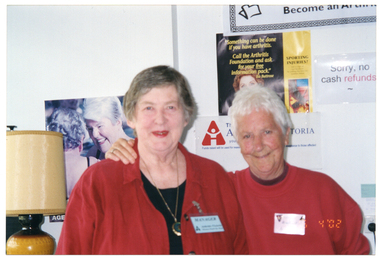 COL medium close up photo of two women inside a shop. One woman has her arm around the other woman's shoulder. They are both wearing red - one, a red shirt on top of a black t-shirt, and the other a red sweatshirt. To the left of the frame, there is a large beige, brown and tan lamp. On the wall behind them, there are some signs and posters. One poster on the left features a large image of two women in a swimming pool. Beside it is a black and yellow poster featuring editor, journalist, and Arthritis Victoria ambassador, Ita Buttrose. The text on this poster says, "Something can be done if you have arthritis. Call the Arthritis Foundation and ask for your free information pack. Ita Buttrose". Below the poster, there is a white Arthritis Victoria sign with a red and white logo and blue text. To the right of this poster and sign is another sign with the text, "Sorry, no cash refunds". In the bottom right corner of the photo image, there is a date stamp in bright orange digits: "16  4 '02".