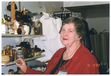 COL medium close up photo of a woman inside a shop. She is wearing a red shirt over a black t-shirt. She is standing next to some wall shelving and has one hand on one of the shelves. On the shelves, there is a range of crockery, glassware, and pottery, including some vases. Alongside the shelving, there is a clothes hanging rack affixed to the wall. Hanging from the rod on coathangers is a range of linen. In the bottom right corner of the photo image, there is a date stamp in bright orange digits: "16  4 '02".
