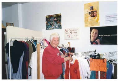 COL medium close up photo of a woman inside a shop. She is wearing a a red sweatshirt. She is standing in front of several racks of clothing, which vary in height. At the front of the shortest rack (to the right of the frame), there is a handwritten sign on an orange piece of paper which says, "LADIES SLACKS $3". On the wall behind the clothing racks, there are some posters and signs. One is a handwritten sign with red text on white paper. It says, "ALL CLOTHING ON THESE 5 RACKS, $3.00 PER PIECE", with some additional text below, "A SUIT OF JACKET & TROUSERS (OR SKIRT) = 2 PIECES". One of the posters is titled, "Make a Splash About Arthritis!", with the sub-heading, "Water exercise is a great way to keep your joints moving." It depicts people doing various types of water exercise and/or therapy in swimming pools or a hot tub. Another poster depicts a middle-aged woman holding a small dumbbell in one hand. Beside the image is the text, "ACTIVE LIVING: GET STRONGER" (partially obscured). In the bottom right corner of the photo image, there is a date stamp in bright orange digits: "16  4 '02".