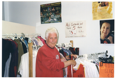 COL medium close up photo of a woman inside a shop. She is wearing a a red sweatshirt. She is standing in front of several racks of clothing, which vary in height. At the front of the shortest rack (to the right of the frame), there is a handwritten sign on an orange piece of paper which says, "LADIES SLACKS $3" (partially obscured). On the wall behind the clothing racks, there are some posters and signs. One is a handwritten sign with red text on white paper. It says, "ALL CLOTHING ON THESE 5 RACKS, $3.00 PER PIECE", with some additional text below, "A SUIT OF JACKET & TROUSERS (OR SKIRT) = 2 PIECES". One of the posters is titled, "Make a Splash About Arthritis!", with the sub-heading, "Water exercise is a great way to keep your joints moving." It depicts people doing various types of water exercise and/or therapy in swimming pools or a hot tub. Another poster depicts a middle-aged woman holding a small dumbbell in one hand. Beside the image is the text, "ACTIVE..." (partially obscured). In the bottom right corner of the photo image, there is a date stamp in bright orange digits: "16  4 '02".