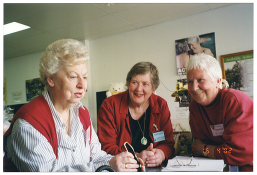 COL medium close up photo of three women leaning on a counter inside a shop. The woman to the left of the frame is holding a pair of spectacles in her hand. The other two women are looking at her, smiling. On the counter, there is some paper or an exercise book, with another pair of spectacles sitting on top. In the background, there are several posters on the wall, and a framed poster with the heading, "Arthritis, Active..." (partially obscured). There is also a white teddy bear with a yellow bow tie (partially obscured), and a vase of flowers with greenery (partially obscured). In the bottom right corner of the photo image, there is a date stamp in bright orange digits: "16  4 '02".