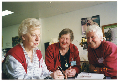 COL medium close up photo of three women leaning on a counter inside a shop. The woman to the left of the frame is holding a pair of spectacles in her hand. The other two women are looking at her, smiling. On the counter, there is some paper or an exercise book, with another pair of spectacles sitting on top. In the background, there are several posters on the wall, and a framed poster with the heading, "Arthritis, Active..." (partially obscured). There is also a white teddy bear with a yellow bow tie (partially obscured), and a vase of flowers with greenery (partially obscured). In the bottom right corner of the photo image, there is a date stamp in bright orange digits: "16  4 '02".
