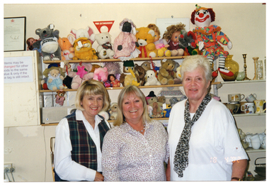 COL medium close up photo of three women inside a shop. The woman on the left is wearing a white shirt with a checked tartan vest; the woman in the centre is wearing a white shirt with small pink flowers; and the woman on the right of the frame is wearing a white t-shirt with a leopard print scarf. They are standing next to one another, in front of some wall shelving. The shelves are filled with soft toys and dolls. To the right of the frame, there are some adjoining shelves which are filled with crockery and bric-a-brac. To the left of the frame, there is a meter box with a typed sign affixed to it. The sign (partially obscured) says, "Items may be changed for other goods to the same value & only if the price tag is still intact". On top of the meter box, there is a framed certificate sitting leaning against the wall. On the bottom right corner of the photo image, there is a date stamp in bright orange digits: "18 4 '02".