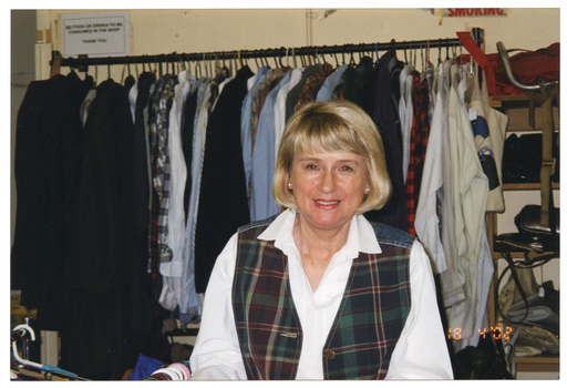 COL medium close up photo of a woman inside a shop. She is wearing a white shirt and a checked tartan vest. She is standing in front of a rack of hanging clothes. Beside the clothes rack are some shelves containing handbags. There are two signs on the wall behind the clothes rack. On the bottom right corner of the photo image, there is a date stamp in bright orange digits: "18 4 '02".