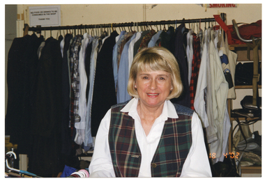 COL medium close up photo of a woman inside a shop. She is wearing a white shirt and a checked tartan vest. She is standing in front of a rack of hanging clothes. Beside the clothes rack are some shelves containing handbags. There are two signs on the wall behind the clothes rack. On the bottom right corner of the photo image, there is a date stamp in bright orange digits: "18 4 '02".