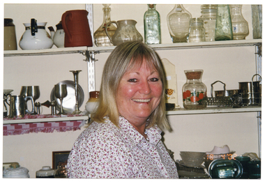 COL medium close up photo of a woman inside a shop. She is wearing a white shirt with small pink flowers. She is standing in three-quarter profile in front of some wall shelving, and smiling. On top of the shelves are various pieces of crockery, glassware, silverware, pewterware and some miscellaneous kitchenware. On the bottom right corner of the photo image, there is a date stamp in bright orange digits: "18 4 '02".