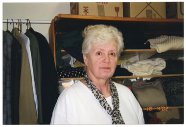 COL medium close up photo of a woman inside a shop. She is wearing a white t-shirt with a leopard print scarf. She is standing in front of a hanging rack of shirts and jackets on coathangers and a bookshelf of folded clothes. There are some boxes on top of the bookcase. On the bottom right corner of the photo image, there is a date stamp in bright orange digits: "18 4 '02".