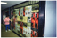 COL photo of a woman inspecting a shop's open glass door and large display window. Both the door and window are completely covered with a range of arthritis-related posters. One poster depitcs a dancer; another poster depicts a woman playing lawn bowls; another poster depicts two people walking along a path in a park; another poster depicts an illustration of a person. There are duplicate posters of each design covering the shop's door and window. The shop appears to be in an arcade.