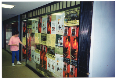 COL photo of a woman inspecting a shop's open glass door and large display window. Both the door and window are completely covered with a range of arthritis-related posters. One poster depitcs a dancer; another poster depicts a woman playing lawn bowls; another poster depicts two people walking along a path in a park; another poster depicts an illustration of a person. There are duplicate posters of each design covering the shop's door and window. The shop appears to be in an arcade.