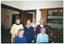 COL photo of a small group of women posing for a photo inside a house. They are sitting or standing behind a table (partially obscured). Behind them is a large display cabinet with plates and silverware. An adjoining room is evident through a large opening in the adjoining wall. Two open doors, one in each room, are also evident.