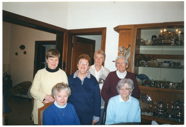 COL photo of a small group of women posing for a photo inside a house. They are sitting or standing behind a table (partially obscured). Behind them is a large display cabinet with plates and silverware. An adjoining room is evident through a large opening in the adjoining wall. Two open doors, one in each room, are also evident.