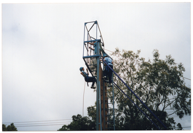 COL photo of four people at the top of a tall metal structure consisting of a ladder which leads to a platform at the top. Two people (one partially obscured) are waiting at the top of the ladder. Another person is standing on the platform. He is looking down towards another person who is descending from the platform via an attached cable. They are all wearing blue tracksuit pants and pale blue polo shirts. There is a large tree in the background.