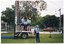COL photo of several teenagers and one adult woman standing on or near a trailer at the base of a tall metal structure with a ladder. The teenagers are all wearing blue tracksuit trousers and pale blue polo shirts. The woman is wearing a white polo shirt and black trousers. Four people are standing on the trailer, while three others, (one partially obscured), look on. One of them has one foot up on the step to the trailer. There is a long cable dangling down from the metal structure. Behind a fence, large trees, school buildings and a red car are evident in the background.