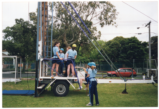 COL photo of several teenagers and one adult woman standing on or near a trailer at the base of a tall metal structure with a ladder. The teenagers are all wearing blue tracksuit trousers and pale blue polo shirts. The woman is wearing a white polo shirt and black trousers. Four people are standing on the trailer, while three others, (one partially obscured), look on. One of them has one foot up on the step to the trailer. There is a long cable dangling down from the metal structure. Behind a fence, large trees, school buildings and a red car are evident in the background.