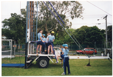 COL photo of several teenagers and one adult woman standing on or near a trailer at the base of a tall metal structure with a ladder. The teenagers are all wearing blue tracksuit trousers and pale blue polo shirts. The woman is wearing a white polo shirt and black trousers. Four people are standing on the trailer, while three others, (one partially obscured), look on. One of them has one foot up on the step to the trailer. There is a long cable dangling down from the metal structure. Behind a fence, large trees, school buildings and a red car are evident in the background.