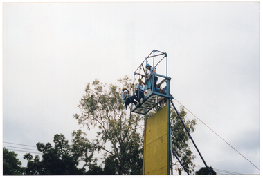 COL photo of a small group of people at the top of a tall metal structure. Most of them are wearing pale blue polo shirts and dark blue tracksuit pants. One man is wearing a blue t-shirt and white trousers. They are all standing on top of a platform. One person is dangling from a cable attached to the metal structure, with her feet resting on the edge of the platform. Another person is climbing from the ladder onto the platform. There are some large trees in the background.