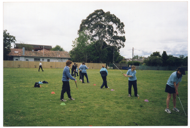 COL photo of a group of people standing on a large, flat grassy area. Most of them are wearing pale blue polo shirts and dark blue tracksuit pants. Two people are wearing a blue jumper or tracksuit jacket on top. Most of the people are standing in a line between field marker cones, which have been set an equal distance apart. They are each holding a golf club, and a few people are practicing their golf swing. In the distance, there is one person walking away at the far end of the grassy area. She is wearing a white shirt and dark trousers. In the background, there are several houses and tall trees behind a wooden fence.