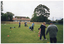 COL photo of a group of people standing on a large, flat grassy area. Most of them are wearing pale blue polo shirts and dark blue tracksuit pants. Two people are wearing a navy blue or black jacket on top, and one woman is wearing a blue and red jumper. Most of the people are standing in a line between field marker cones, which have been set an equal distance apart. A few people are crouching down on the ground. Three people are each holding a golf club and practicing their golf swing. Three of the people are assisting or instructing the others. In the background, there are several houses and tall trees behind a wooden fence.