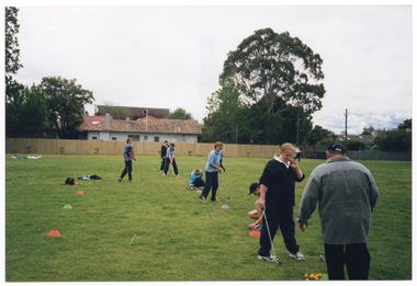 COL photo of a group of people standing on a large, flat grassy area. Most of them are wearing pale blue polo shirts and dark blue tracksuit pants. Two people are wearing a navy blue or black jacket on top, and one woman is wearing a blue and red jumper. Most of the people are standing in a line between field marker cones, which have been set an equal distance apart. A few people are crouching down on the ground. Three people are each holding a golf club and practicing their golf swing. Three of the people are assisting or instructing the others. In the background, there are several houses and tall trees behind a wooden fence.