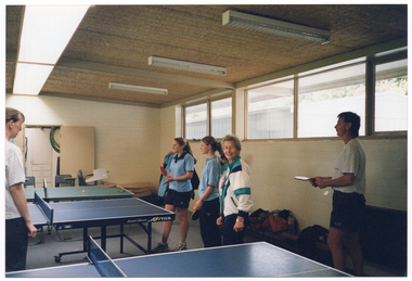 COL photo of several people stand arounding some table tennis tables. Some people are wearing pale blue polo shirts, some are wearing white t-shirts, one woman is wearing a navy, jade, and white tracksuit jacket, and another person (partially obscured) is putting on a blue and navy jacket. They are all wearing either navy shorts or navy tracksuit pants. They are inside a white brick building. A few of them are holding a tennis table racket (also known as a bat or paddle) in their hand. A storage room is evident through an open doorway. There is a view of another building and some trees through the high curtained windows.