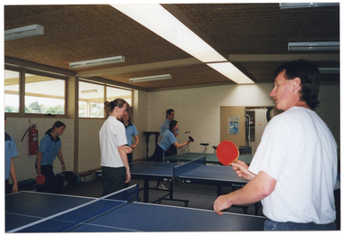 COL photo of a group of people playing table tennis on three table tennis tables. Several people are wearing pale blue polo shirts, two people are wearing white t-shirts, and one person is wearing a blue collared shirt. They are all wearing navy or black trousers or tracksuit pants. They are inside a white brick building. A storage room is evident through an open doorway. There is a view of some trees through the high curtained windows.