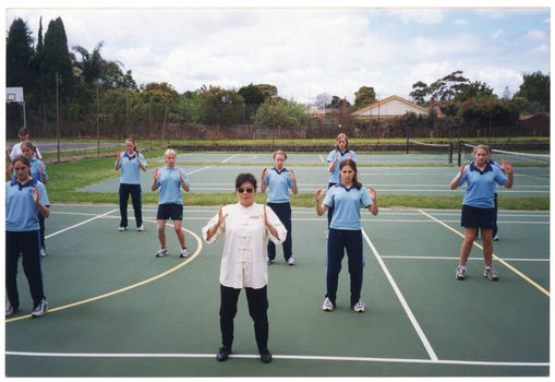 COL photo of a woman leading a group of students in an exercise class. The students are dressed in their sports uniform, consisting of a blue polo top and either navy blue shorts or track suit pants. The instructor is wearing a cream traditional Chinese shirt with a mandarin collar and black trousers. They are standing on an outdoor tennis court in rows, spaced well apart from one another. The instructor is standing in the front row, facing the camera, with her back to the students. There are two more tennis courts in the background, and the entire court area is enclosed by a tall cyclone fence.
